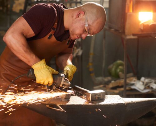 Concentrated busy young workman in safety goggles and leather apron polishing metal with grinder on anvil in workshop