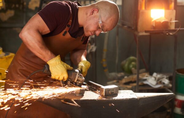 Concentrated busy young workman in safety goggles and leather apron polishing metal with grinder on anvil in workshop